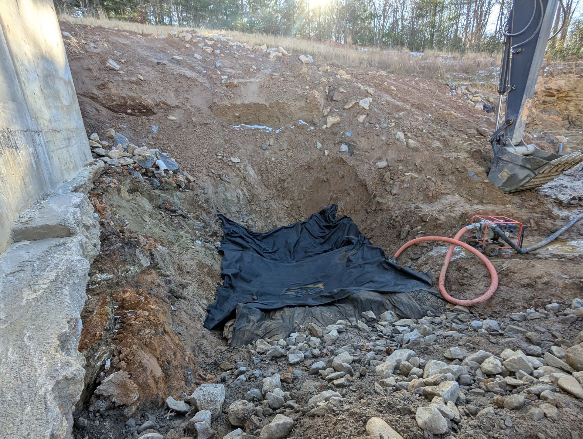 Excavation at a construction site; black tarp covers a hole near a concrete structure and dirt hill.