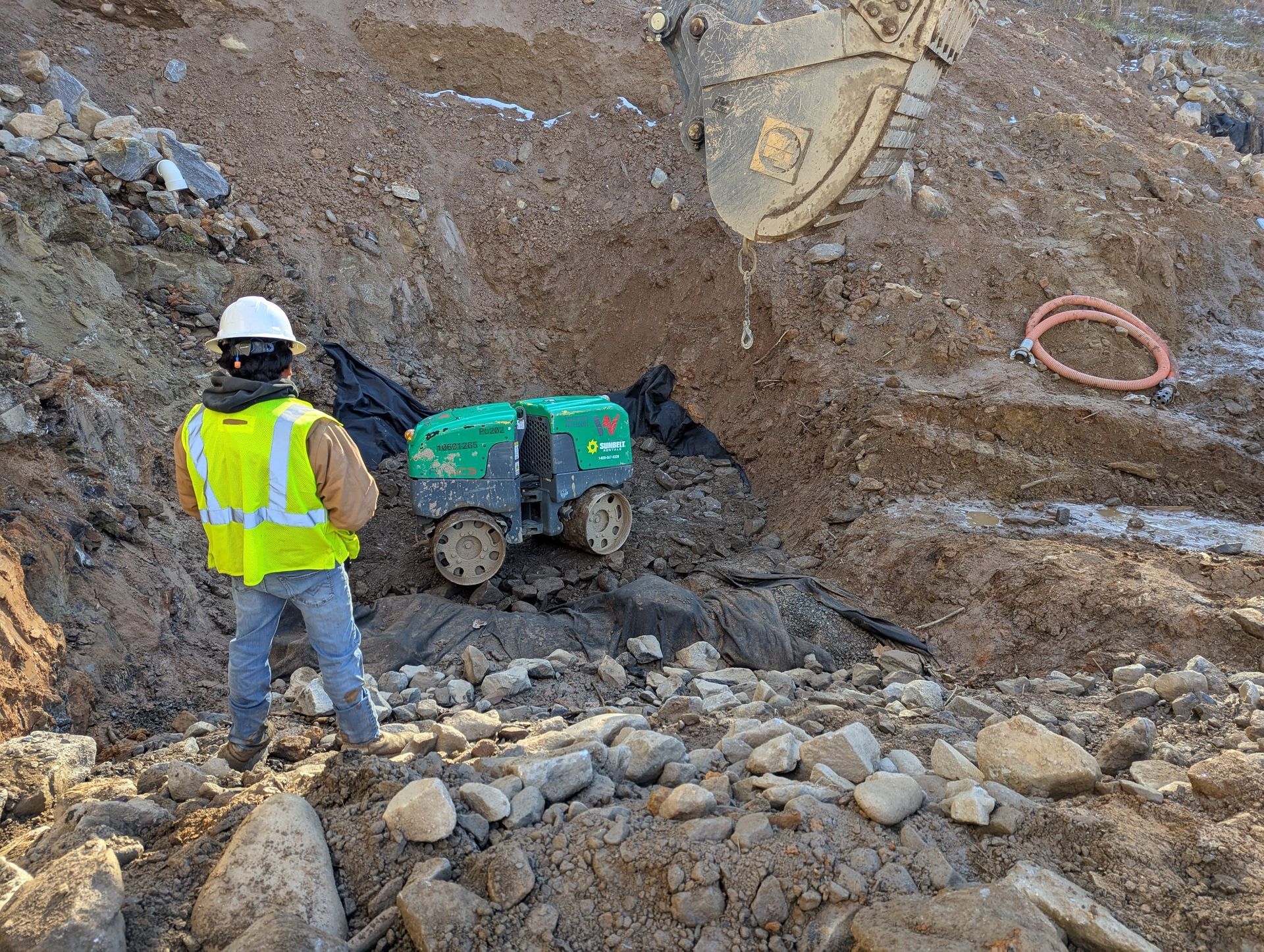 Man in vest watches robot near excavator in dirt pit.