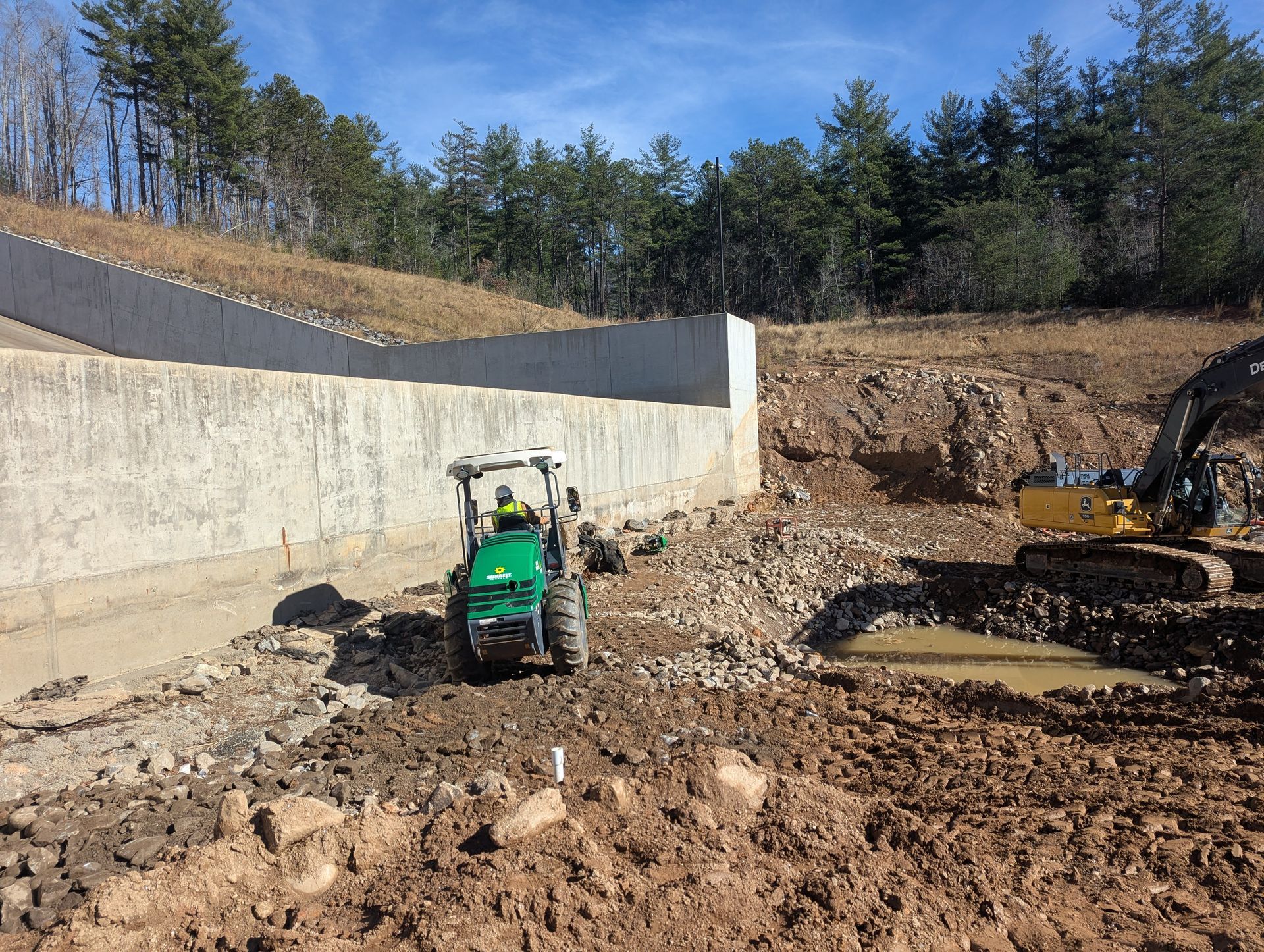 Construction site: Green tractor and yellow excavator working near a concrete retaining wall.