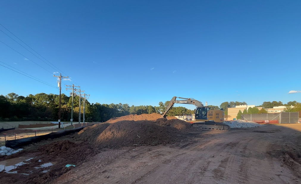 Construction site with excavator on a pile of dirt under a blue sky, power lines and trees in the background.