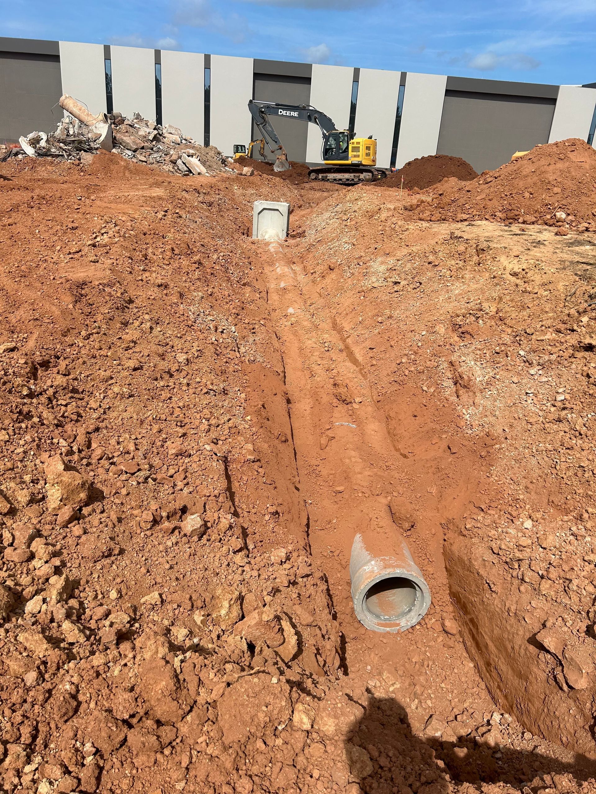 Trench dug in red soil with concrete pipes; excavator and building in the background, construction site.