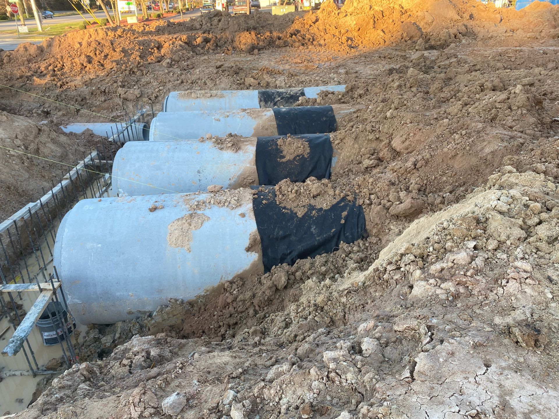 Concrete pipes buried in a trench, construction site. Black tarp covers some pipes, brown soil surrounds.