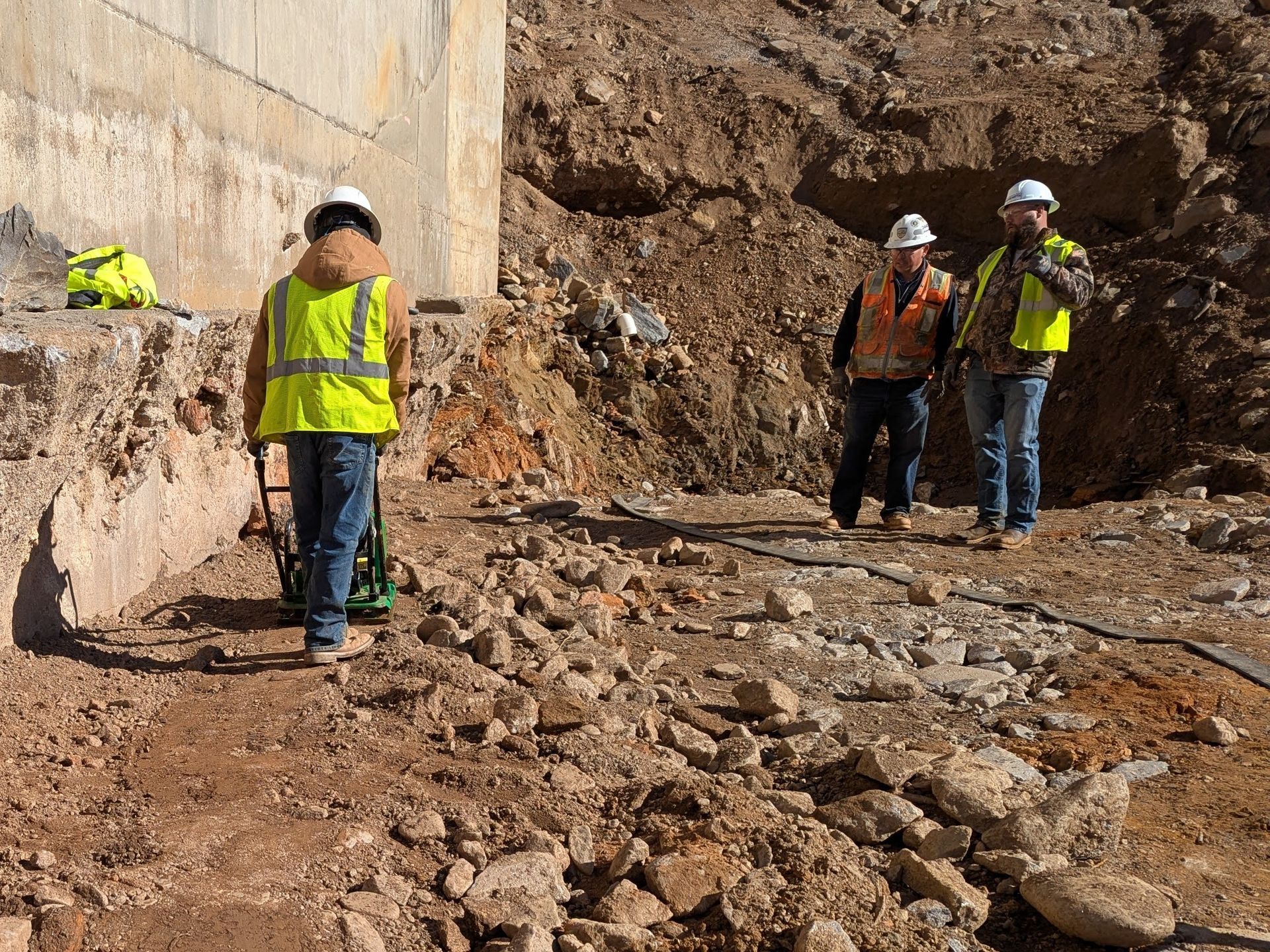 Three construction workers surveying a rocky excavation site near a concrete structure.
