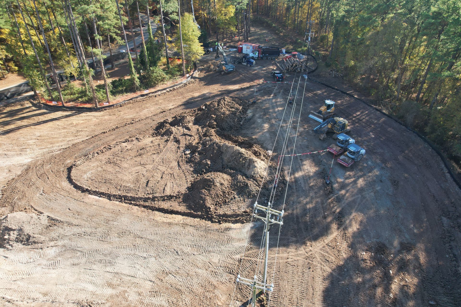 Construction site with heavy machinery, power lines, and cleared land surrounded by trees.