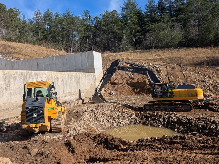 Two yellow construction vehicles excavating at a site with a concrete wall and hillside backdrop.