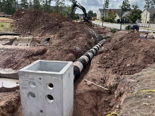 Construction site with a concrete drain, pipes, and excavator; dirt pile, and homes in the background.
