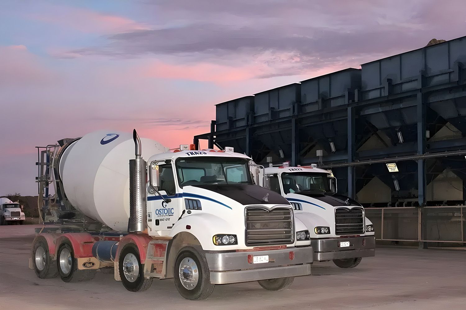 Two White Cement Mixer Trucks Parked In Front Of Industrial Storage Silos