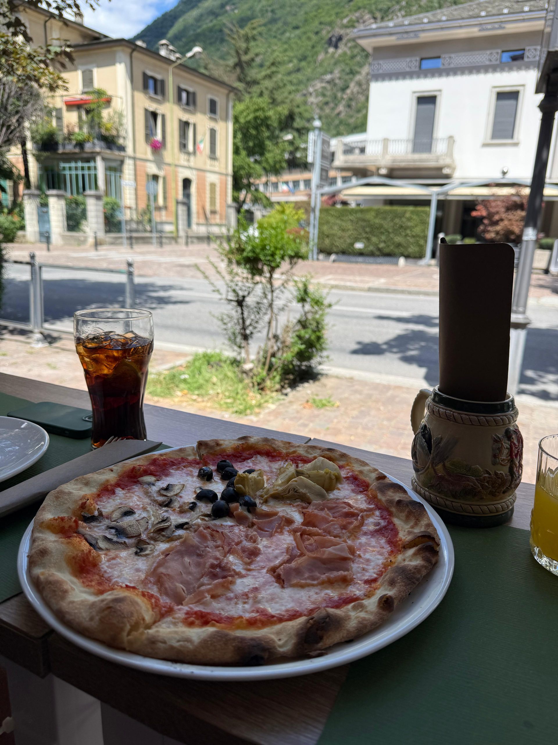 Pizza on a plate with a drink and building in the background.
