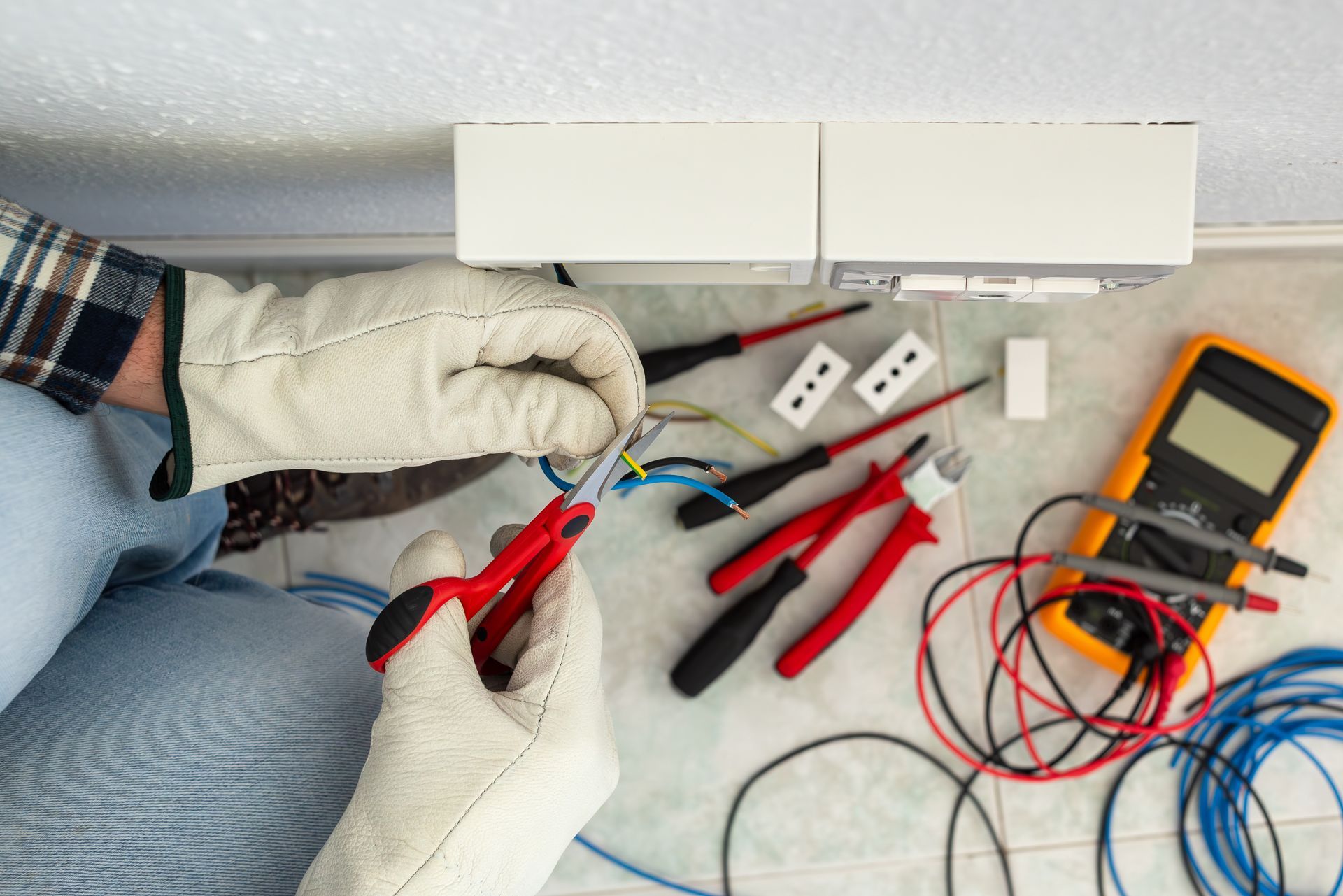 Person wearing gloves cutting wires with scissors near electrical outlet and tools.