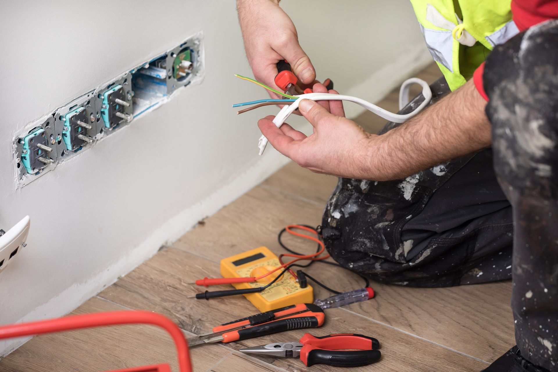 Electrician wiring outlets on a wall, tools on floor, work clothes.