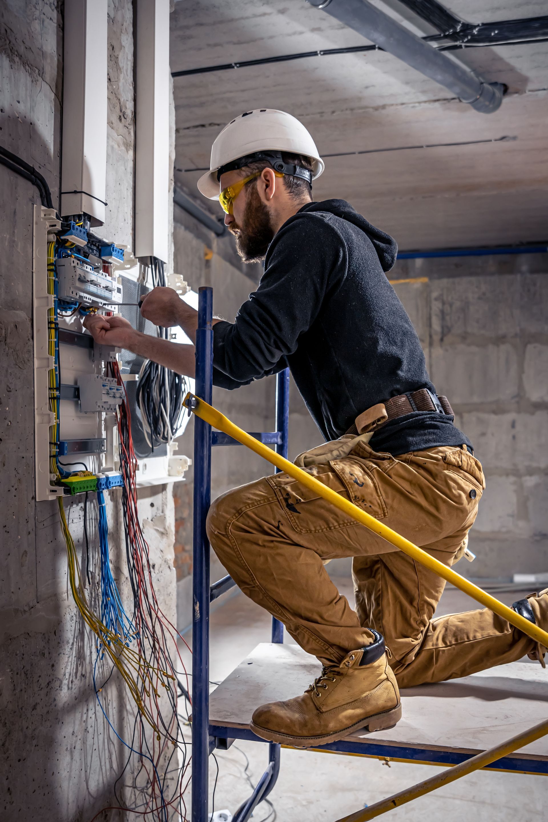Electrician working on electrical panel, wearing safety gear, inside a building.