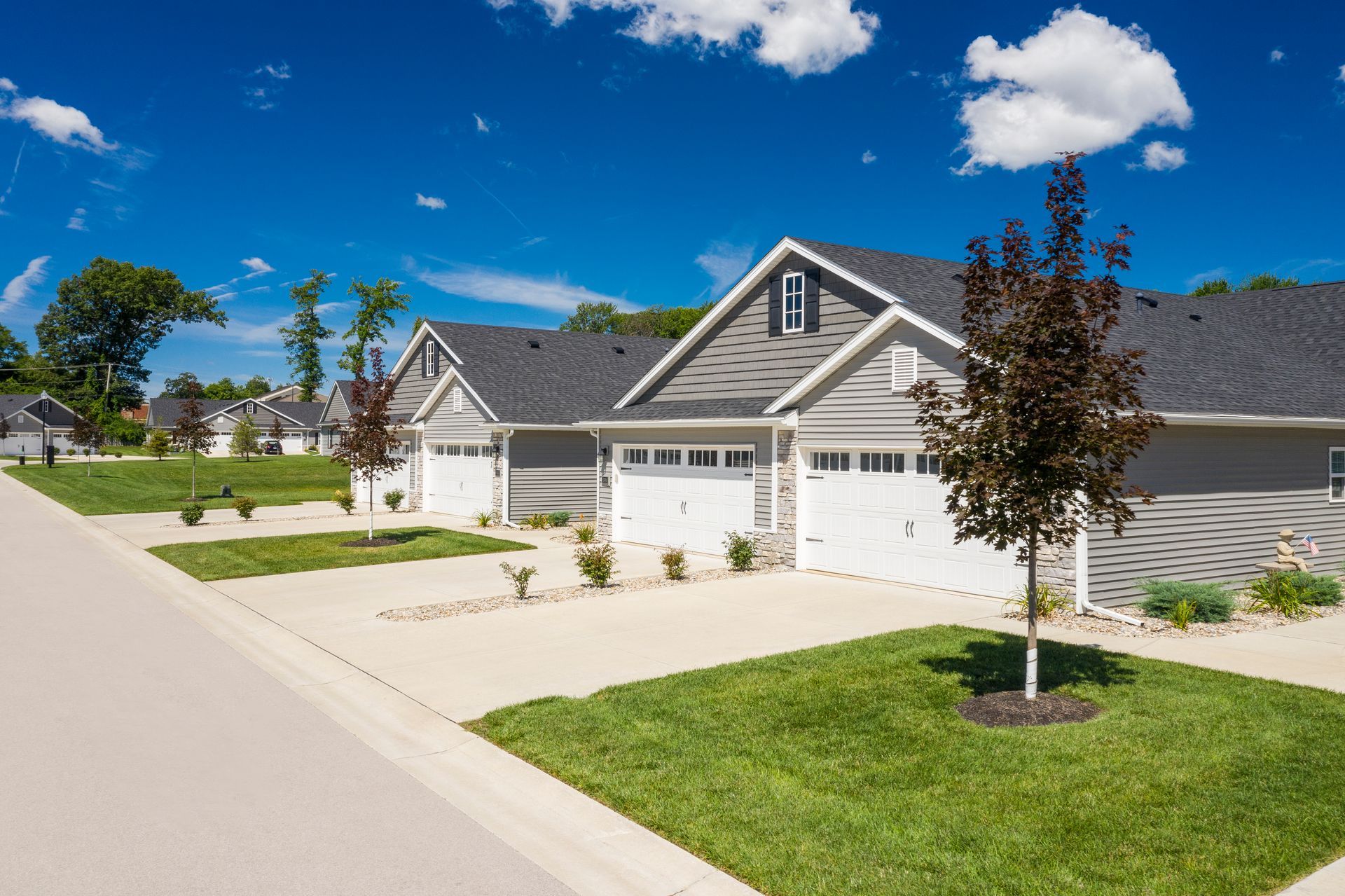 Row of gray houses with white garage doors, green lawns, and a blue sky.