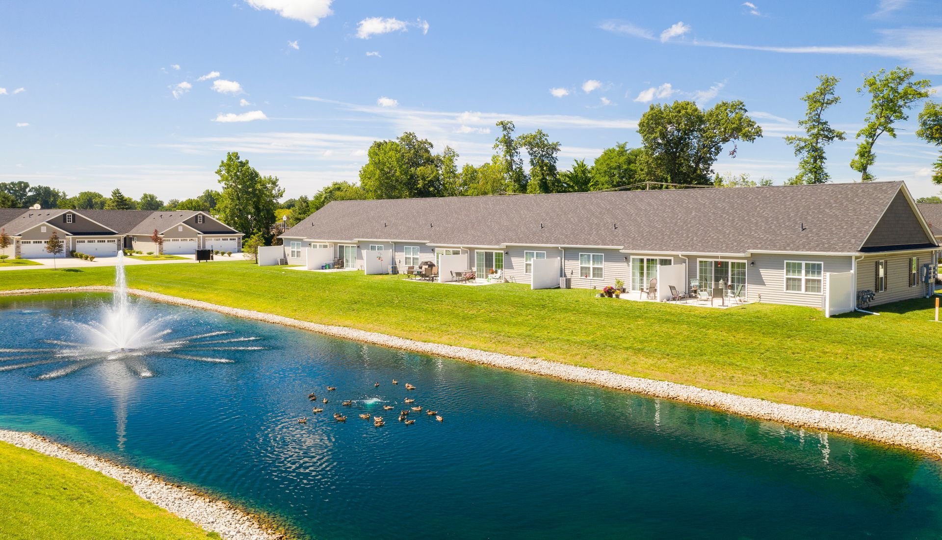Long row of houses with gray roofs beside a pond with a fountain, blue sky.