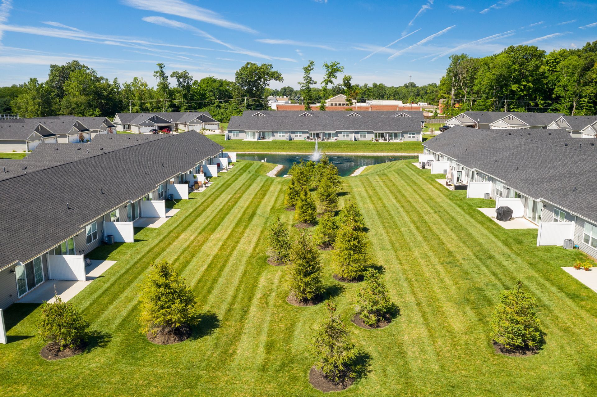 Aerial view of townhouses, central pond, manicured lawns, trees, blue sky.