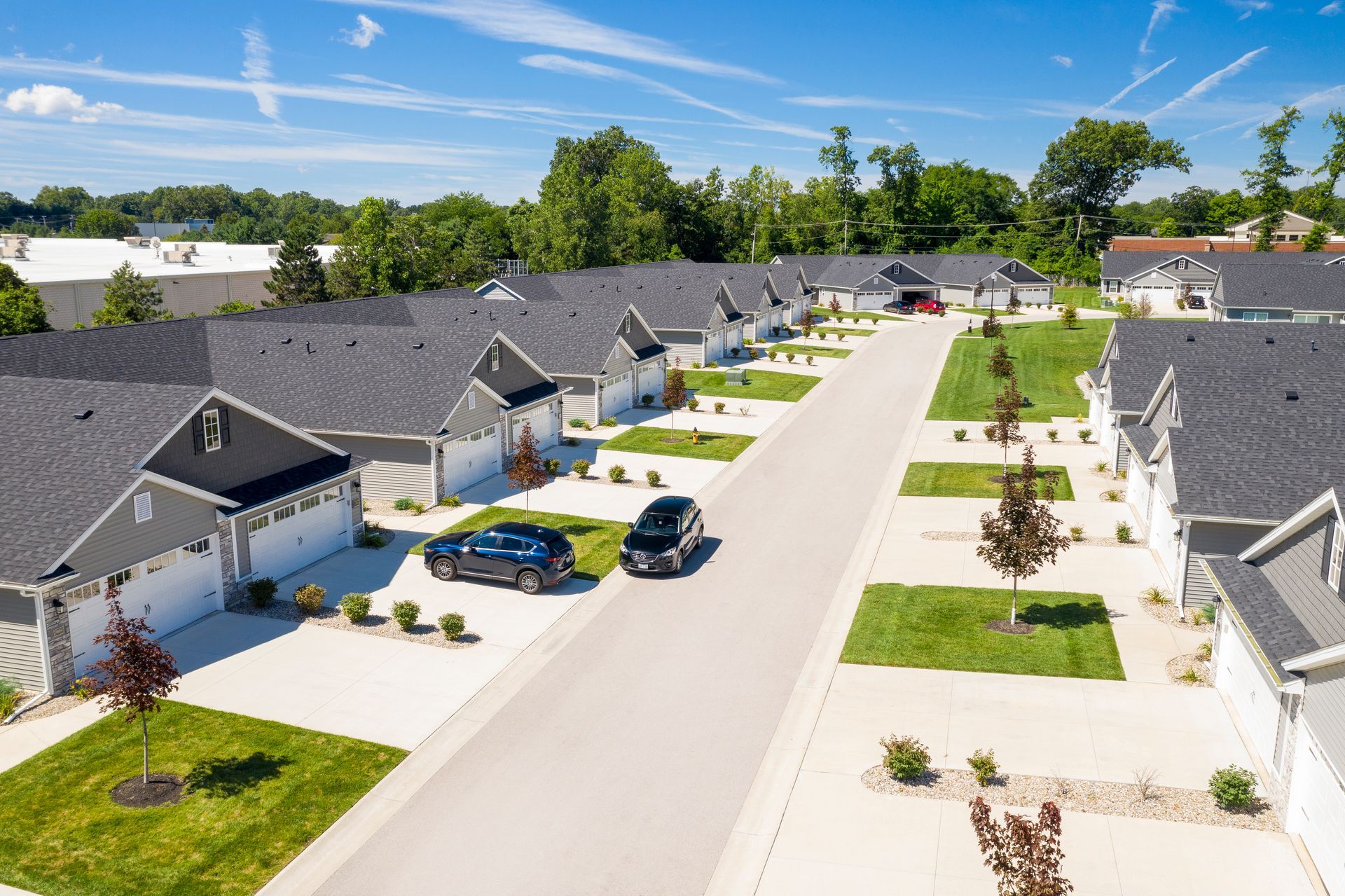 Aerial view of a street lined with modern houses, gray roofs, and green lawns, cars parked. Sunny day.