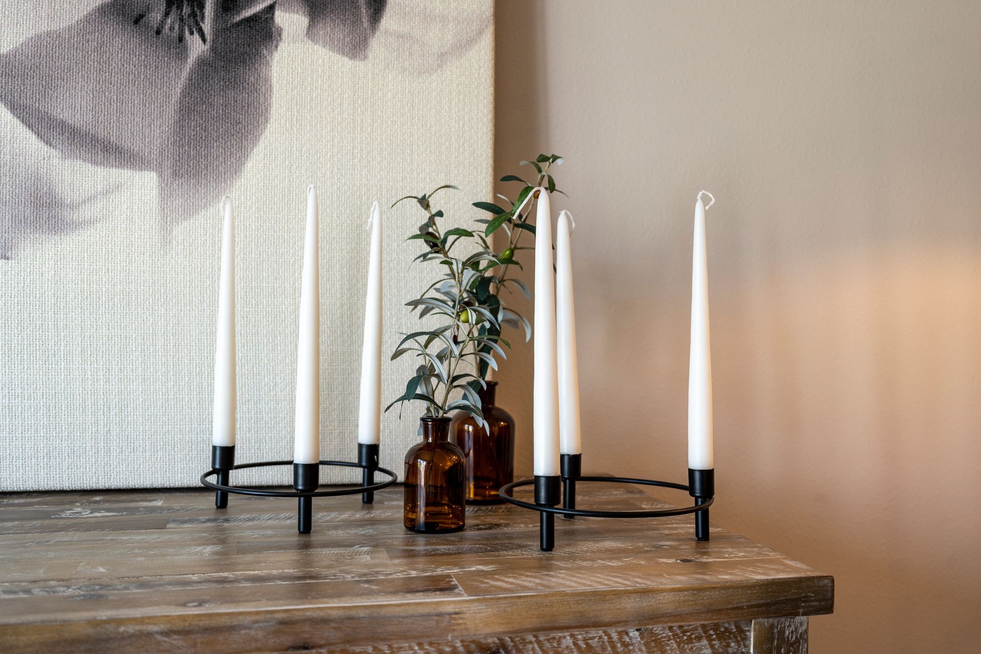 Candle holder with tall white candles on a wooden surface, with a painting in the background.