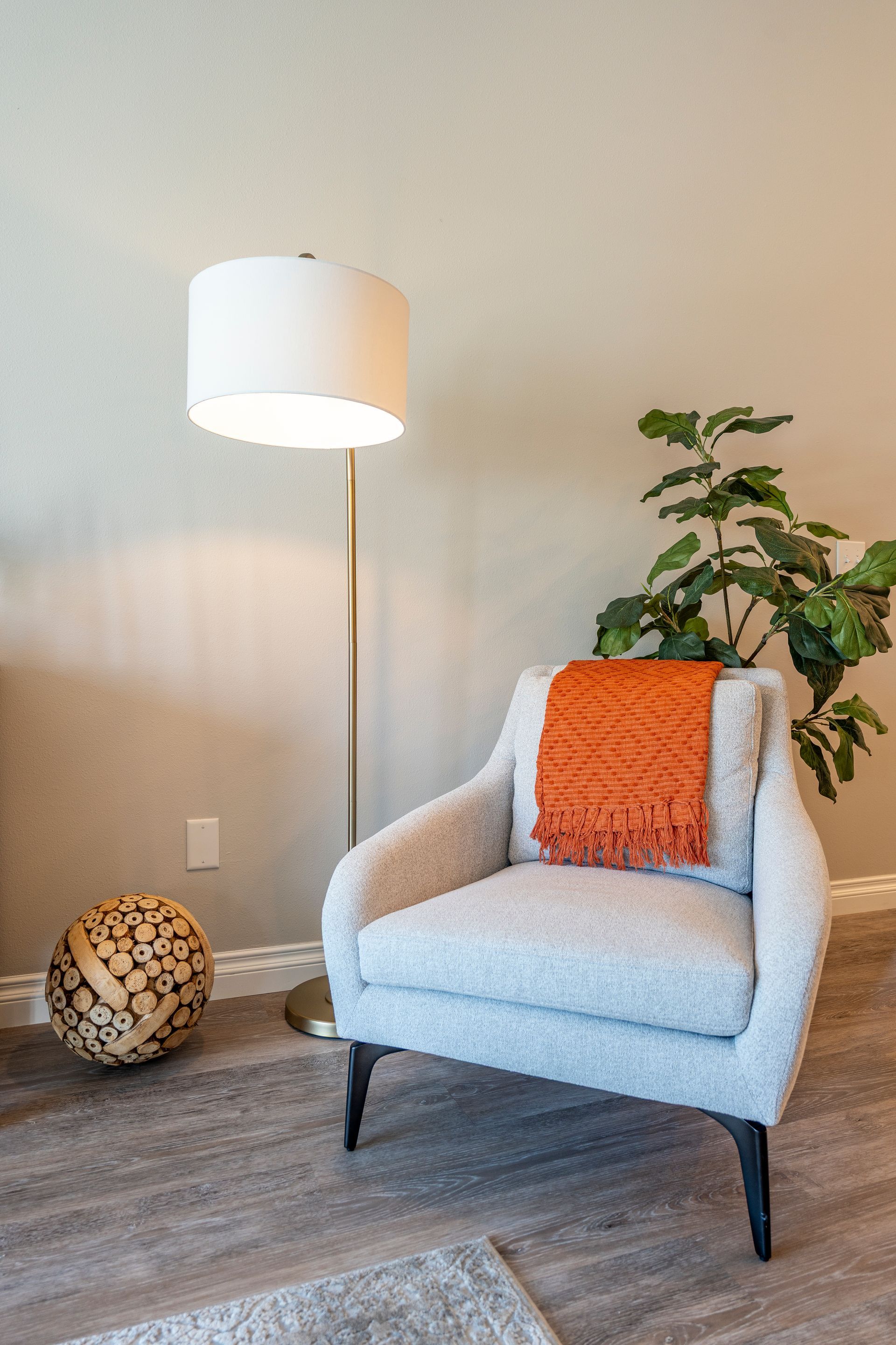Cozy reading nook: Light gray armchair with orange blanket, floor lamp, and leafy plant against a beige wall.