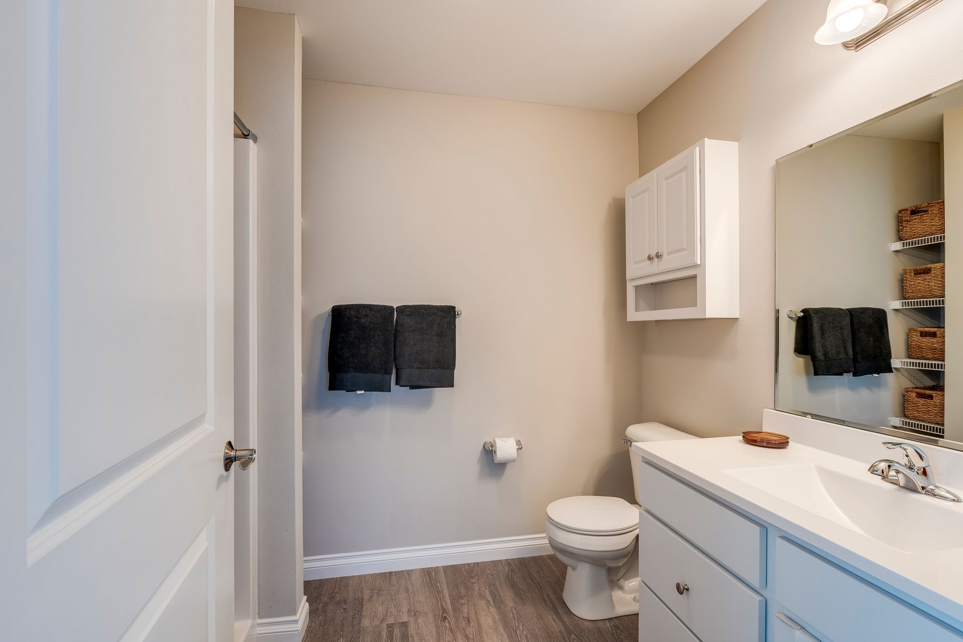 Bathroom with white vanity, toilet, and cabinet. Gray towels hang on the wall, and there’s a large mirror and shelves.