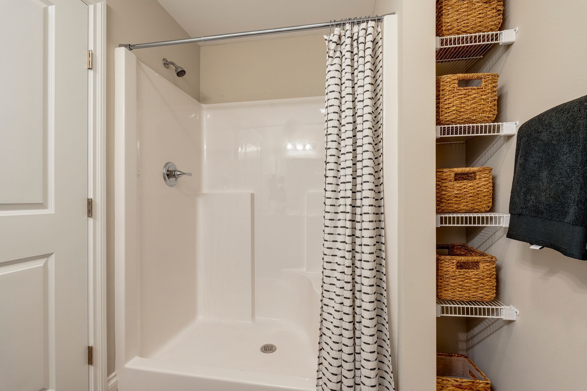White shower with patterned curtain. Wire shelves with woven baskets. A towel hangs on the side.