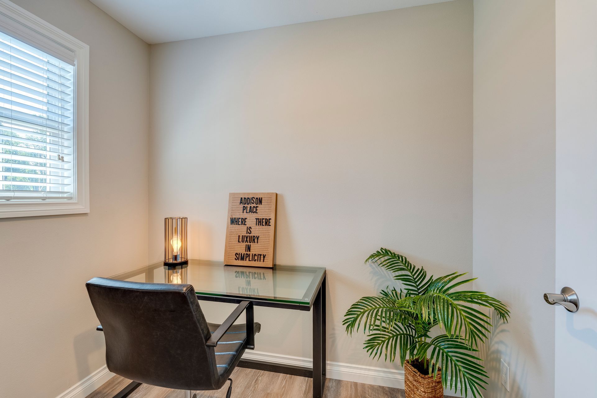 Home office with a glass desk, chair, lamp, plant, and sign near a window and door.