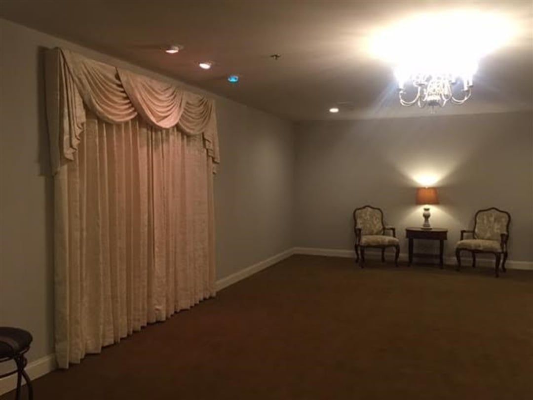 Empty room with ornate drapes, two chairs, side table, and chandelier.