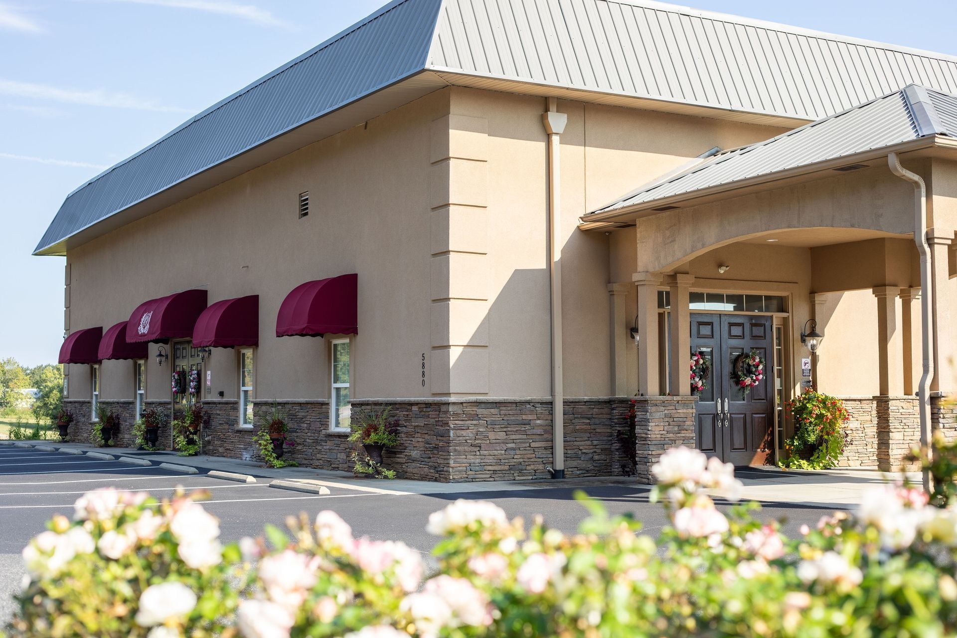 Beige building with burgundy awnings and a stone facade, fronted by roses and a parking lot.