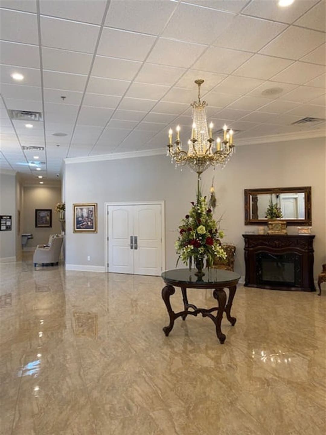 Elegant lobby with chandelier, table, fireplace, and double doors. Shiny floors and neutral walls.