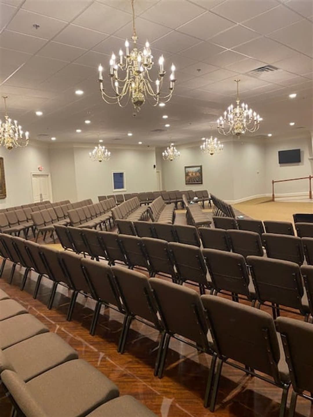Rows of brown chairs face a stage in a large, empty room with chandeliers.
