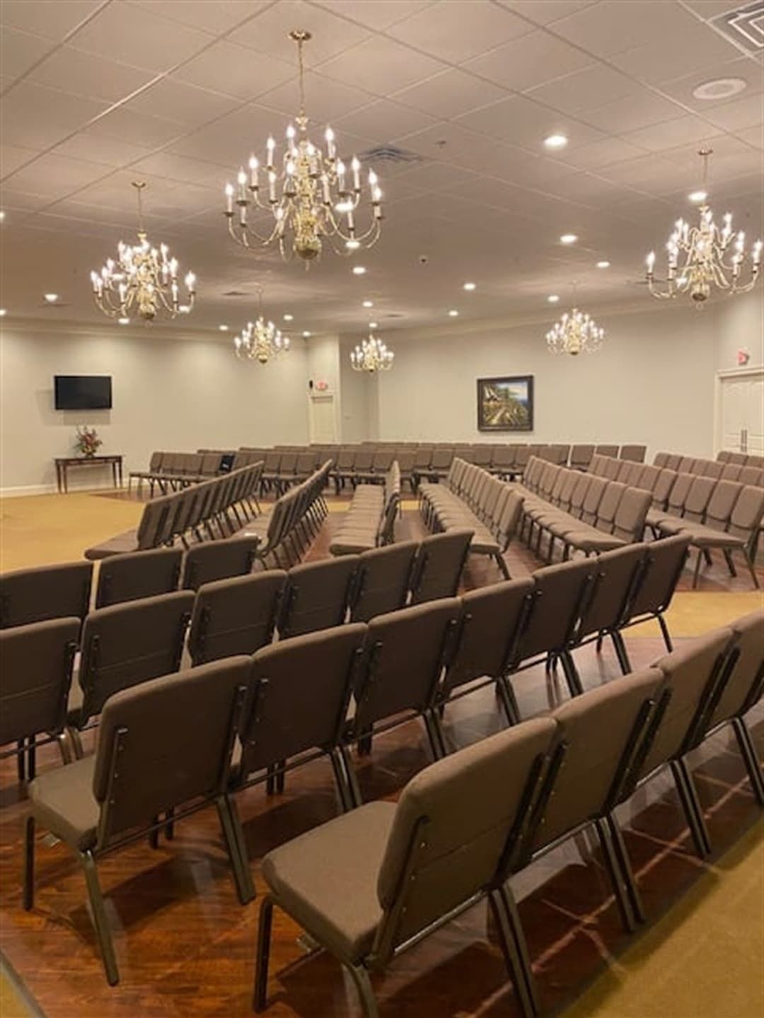 Rows of brown chairs face the front of a large, well-lit room with chandeliers.