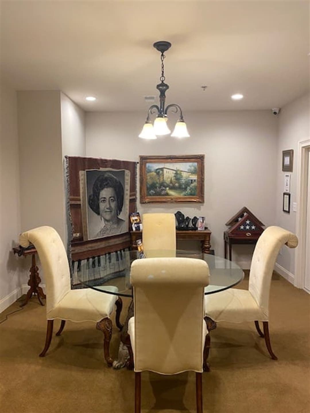Dining room with round glass table, four cream chairs, piano portrait, landscape painting, and chandelier.