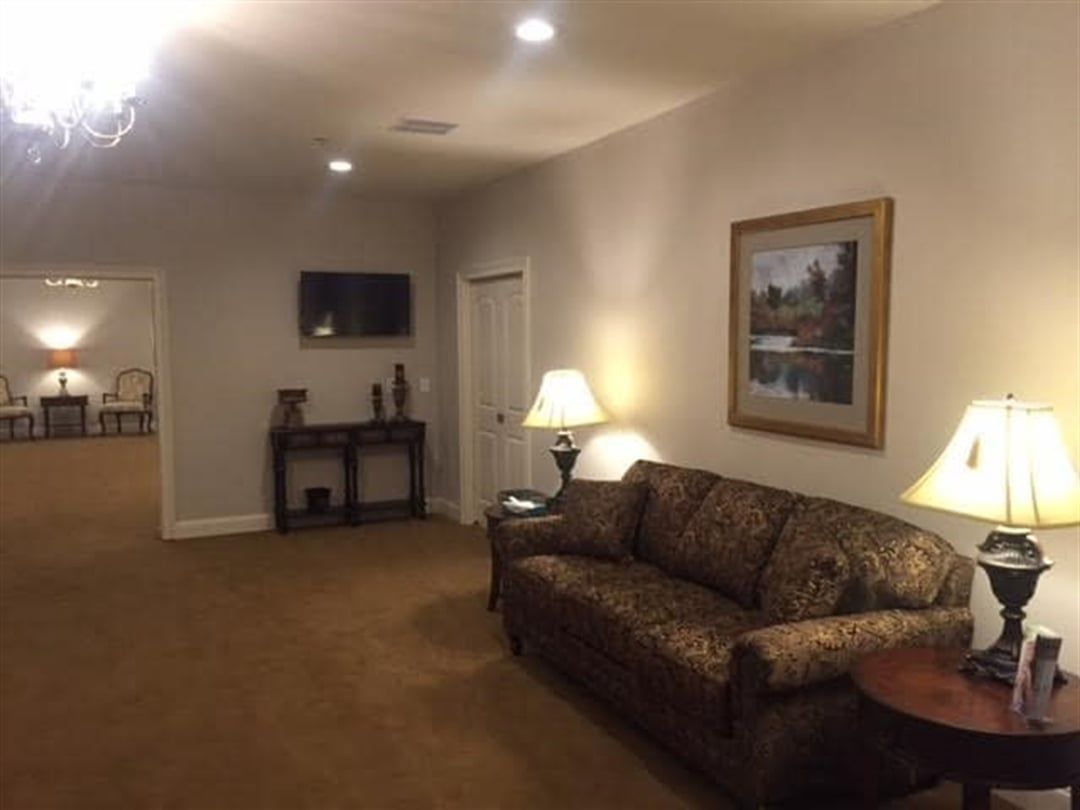 A waiting room with a brown sofa, lamps, and a painting. Another room with chairs is visible through an opening.