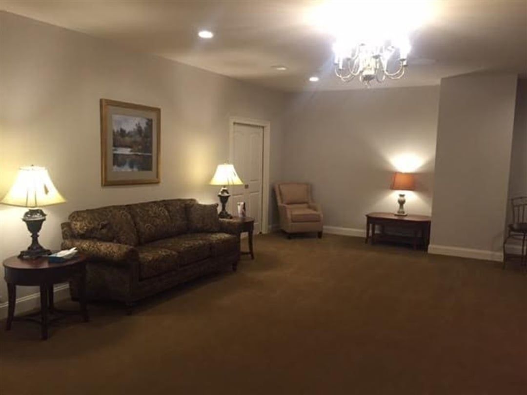 A waiting room with neutral walls, brown carpet, sofas, lamps, and a small table.