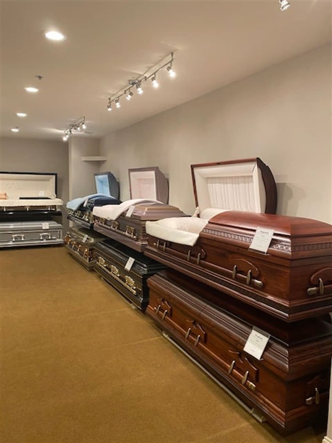 A funeral home showroom with multiple caskets on display. Wooden and metallic caskets of varying colors.