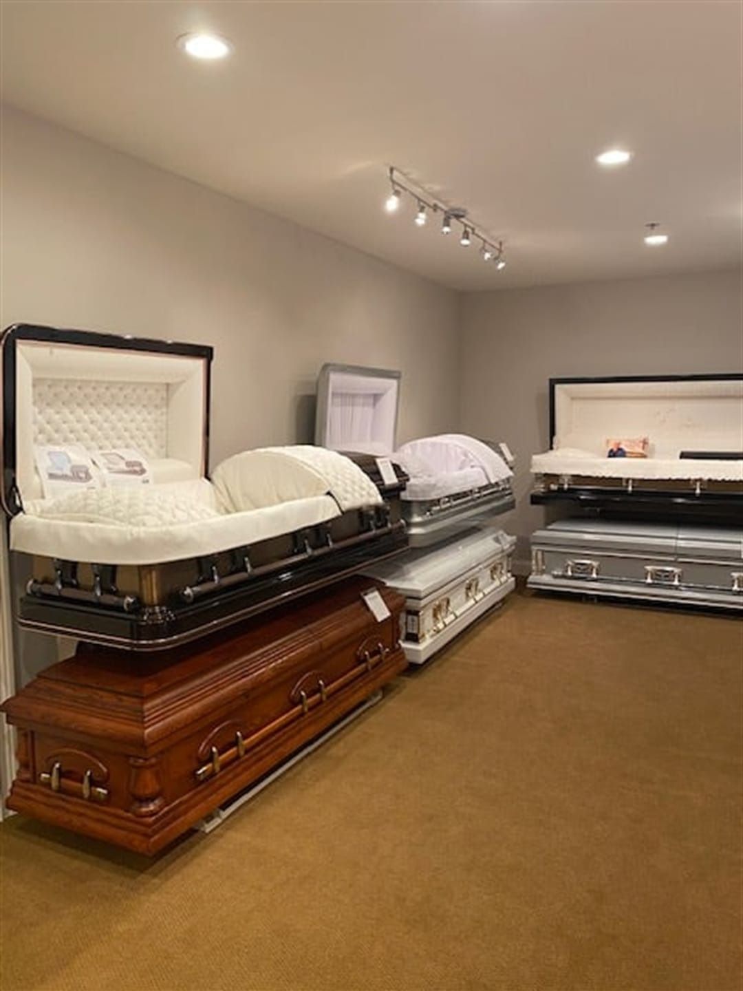 A funeral home showroom displays several caskets, including open ones with interior padding. Brown carpet and neutral walls.