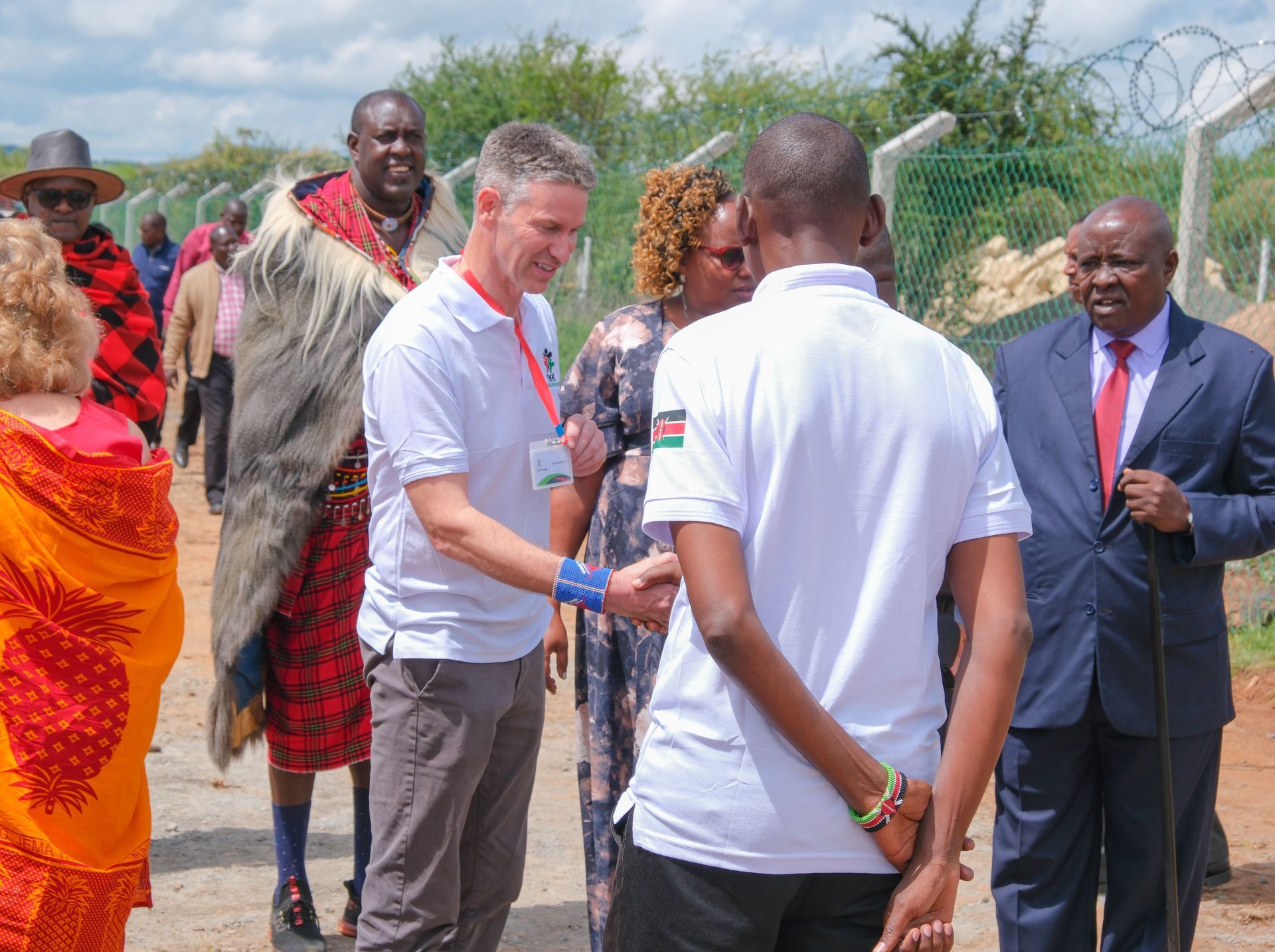 Group of people shaking hands outdoors near a fence. Some wearing traditional Maasai clothing.