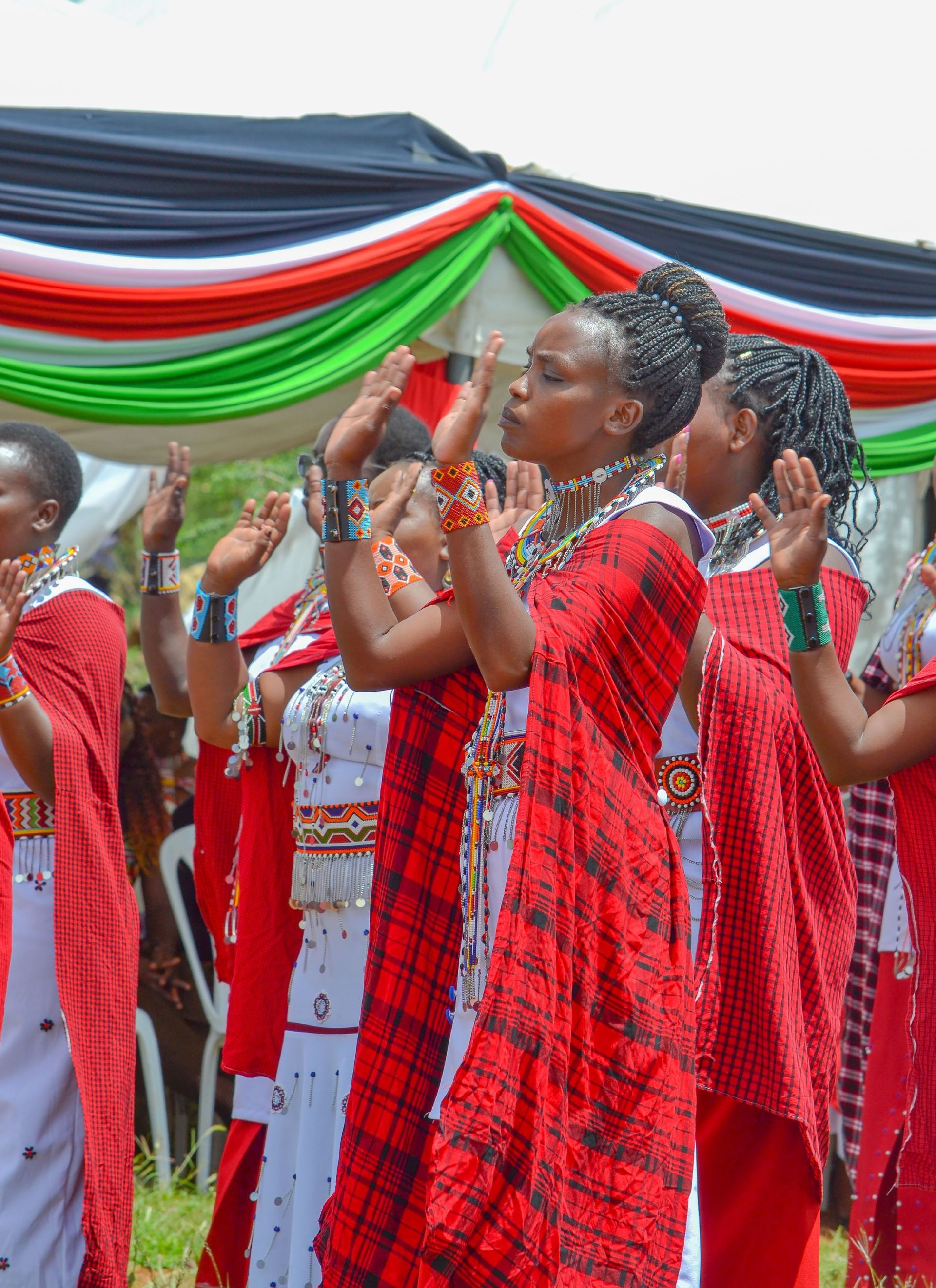 Women in red patterned clothing perform traditional dance with hands raised, under colorful decorations.