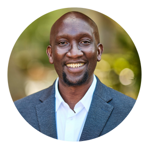 Smiling Black man in a gray suit, white shirt, outdoors with blurred greenery in the background.