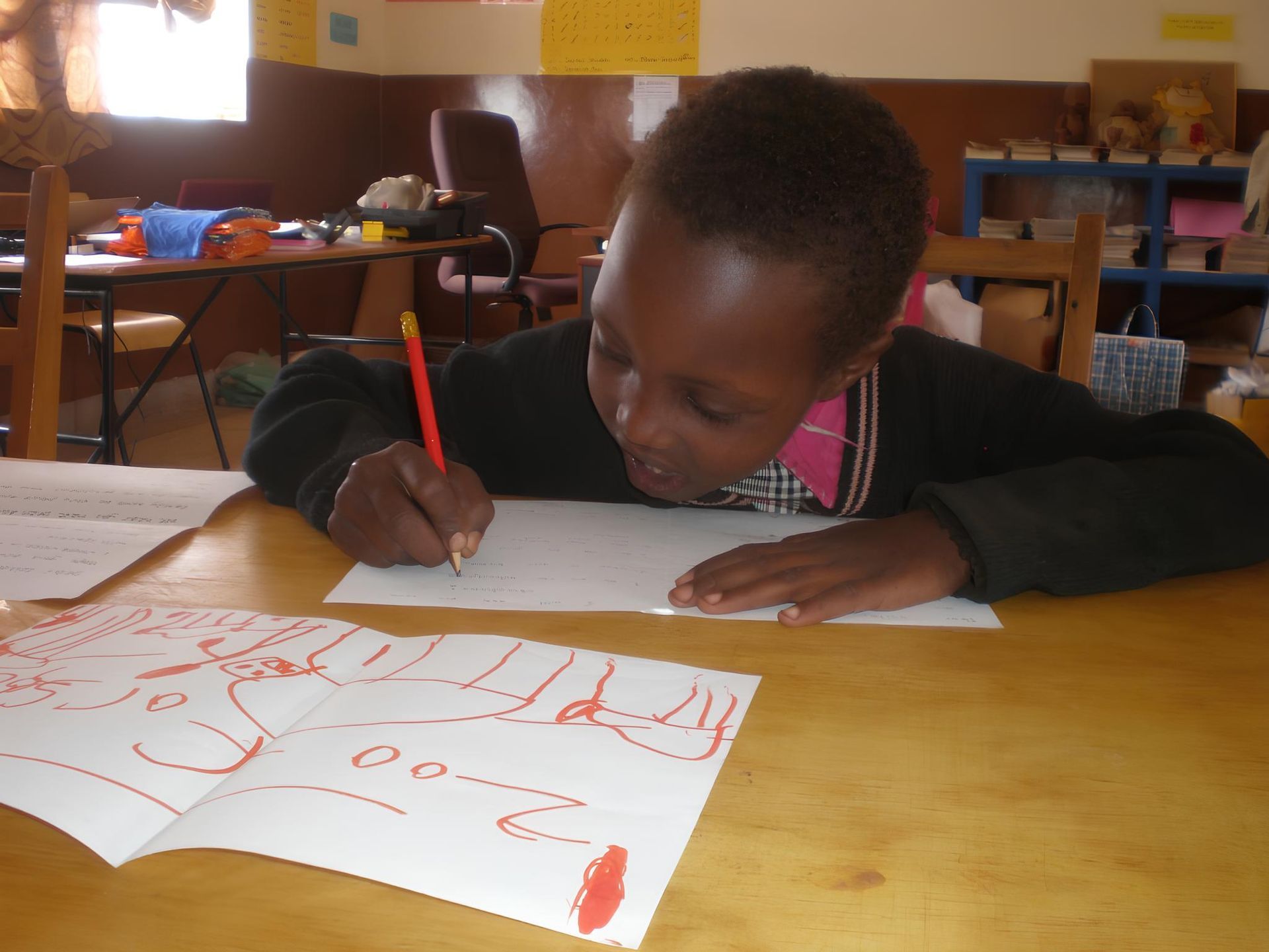 A young Black child writing with a red pencil at a wooden desk, book open, in a classroom.
