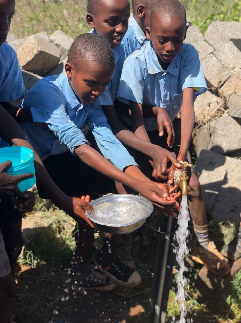Children washing hands at a water tap outdoors. They are in blue uniforms.