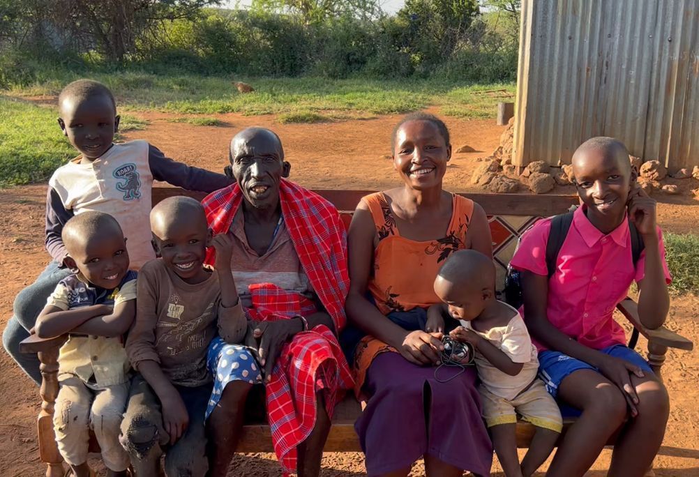 Family sits on a bench outdoors; smiling and dressed in colorful clothes, with children and an elder.