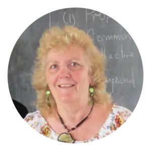 Woman with blonde hair, smiling, wearing earrings and necklace, in front of a chalkboard with writing.