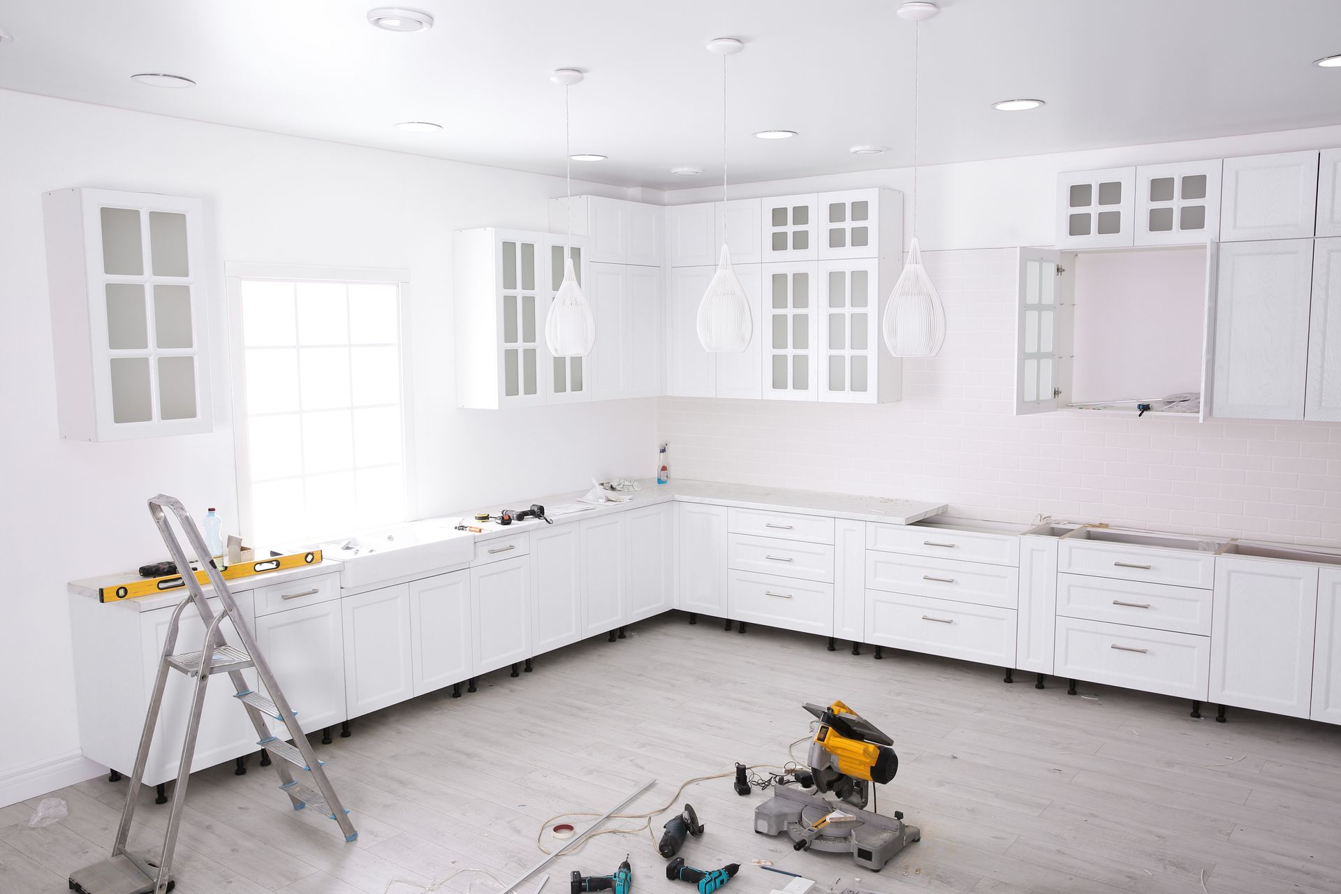 White kitchen cabinets being installed in a room with a window, step ladder, and tools.