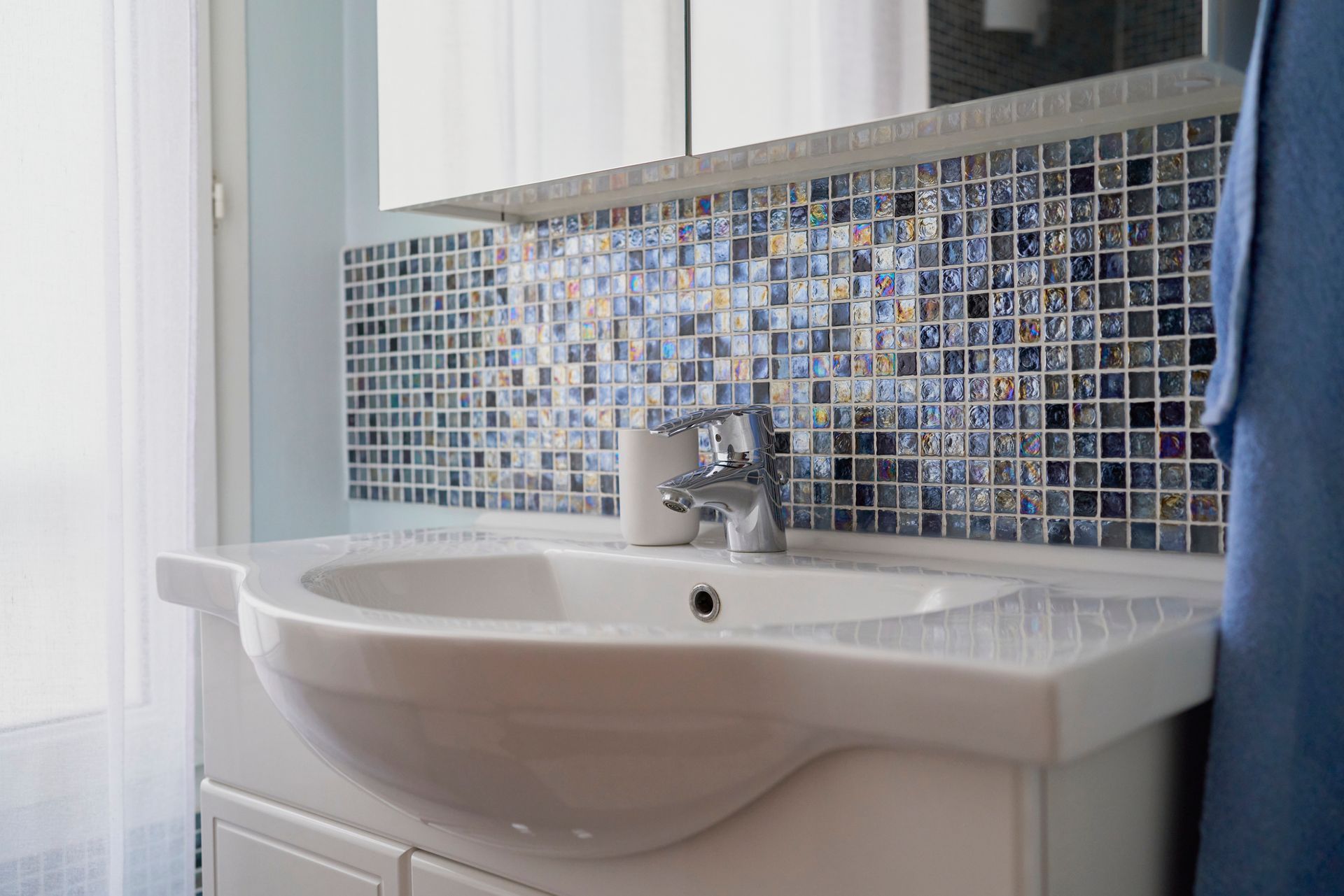Sink with a metal faucet in a bathroom with a multi-colored tile wall next to a window with a curtain.