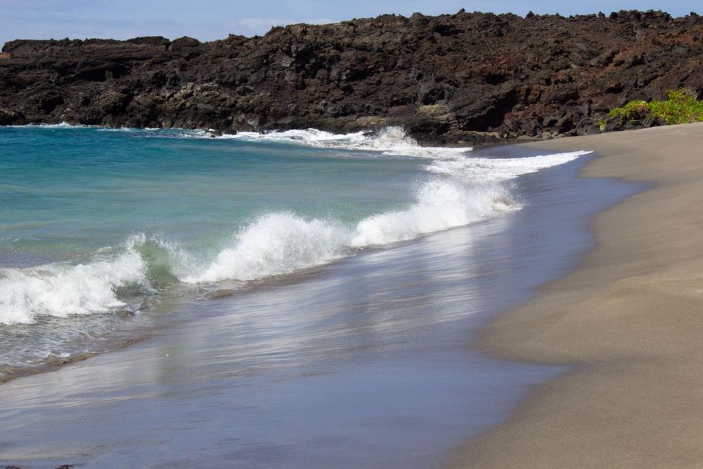 Waves crashing onto a black sand beach with dark lava rock cliffs in the background; blue-green water.
