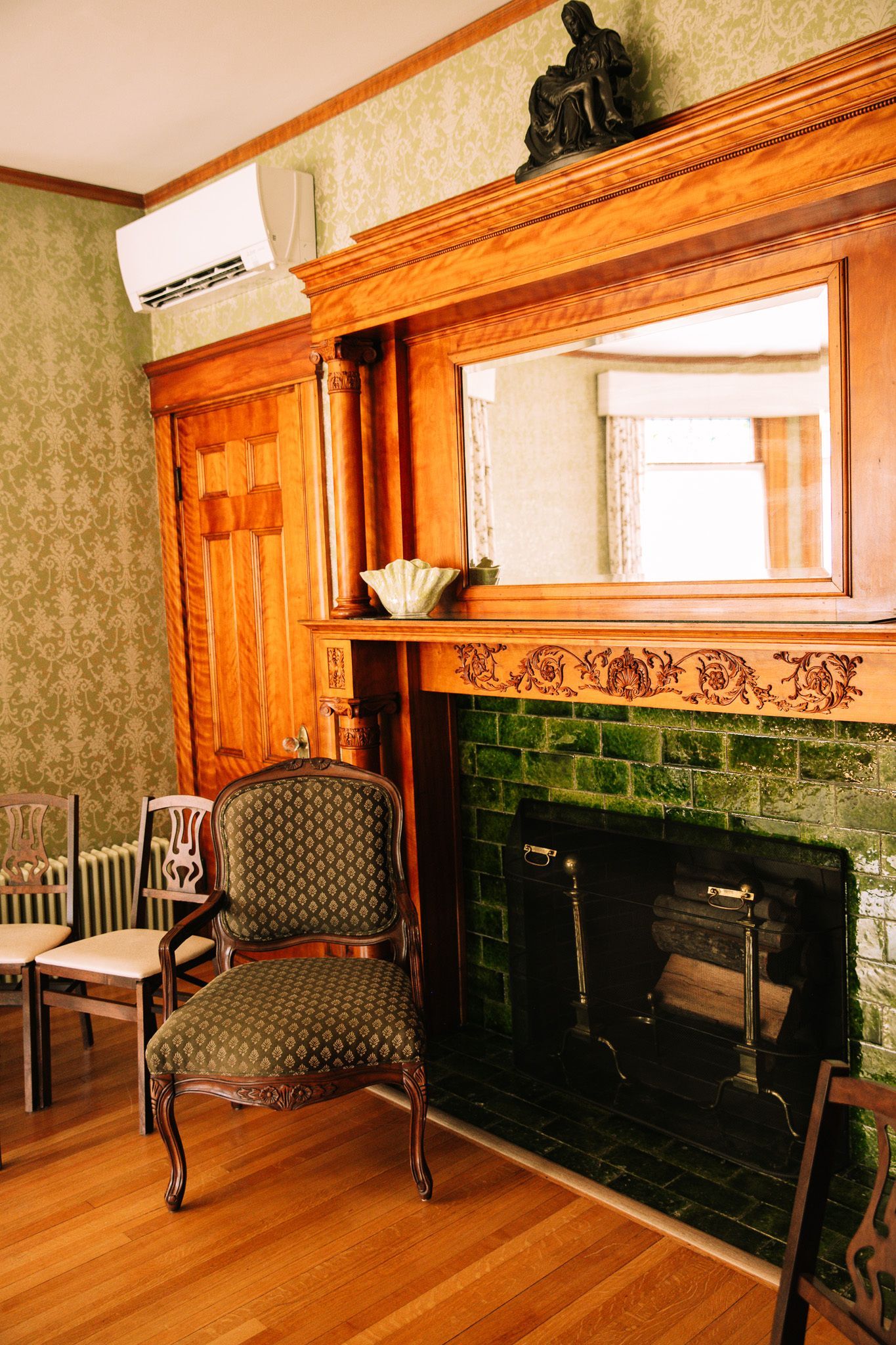 Ornate fireplace with green tiles, wood paneling, mirror, and antique chair in a room with patterned wallpaper.