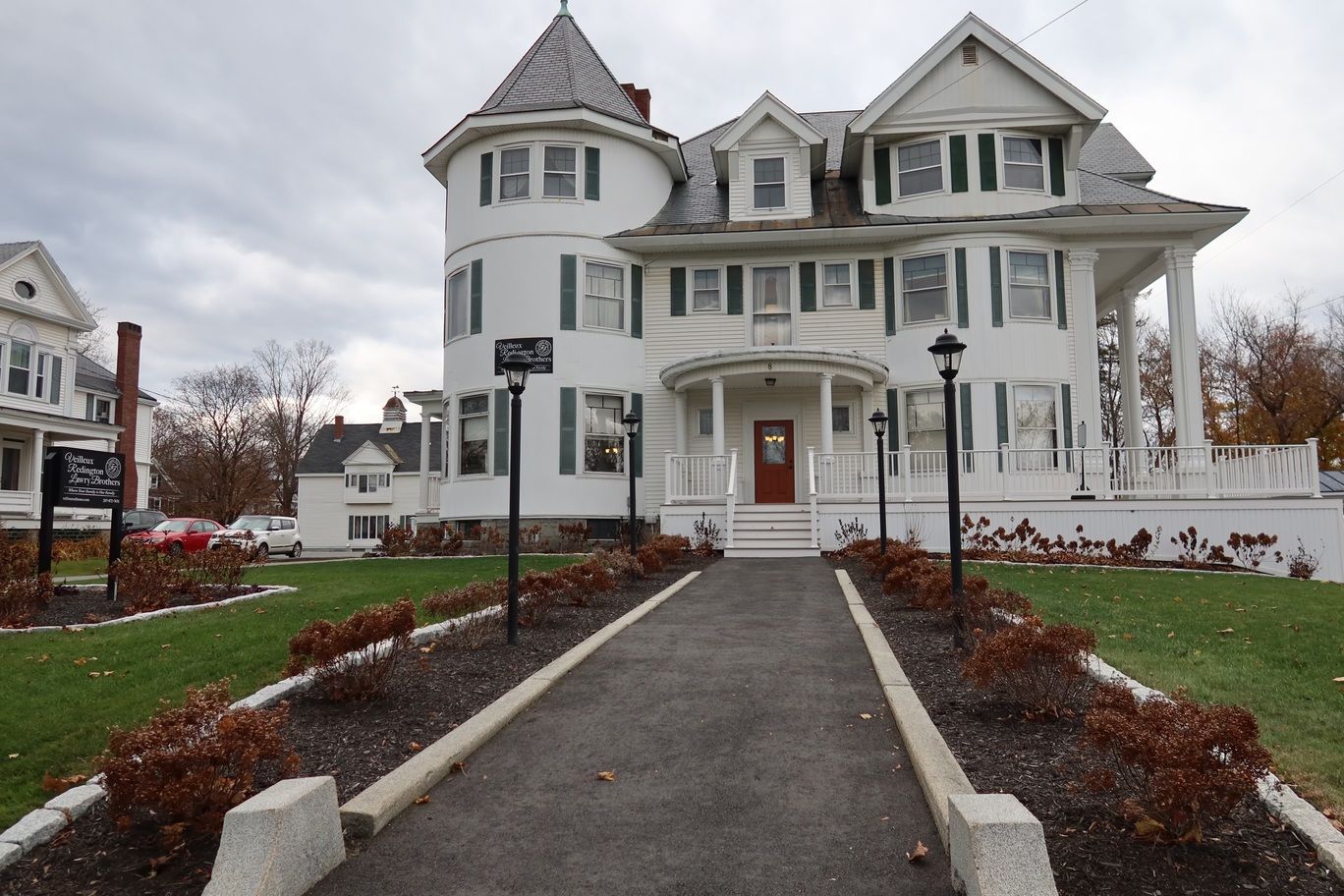 White Victorian building with a turret and dark green shutters, with a walkway leading to a red door.