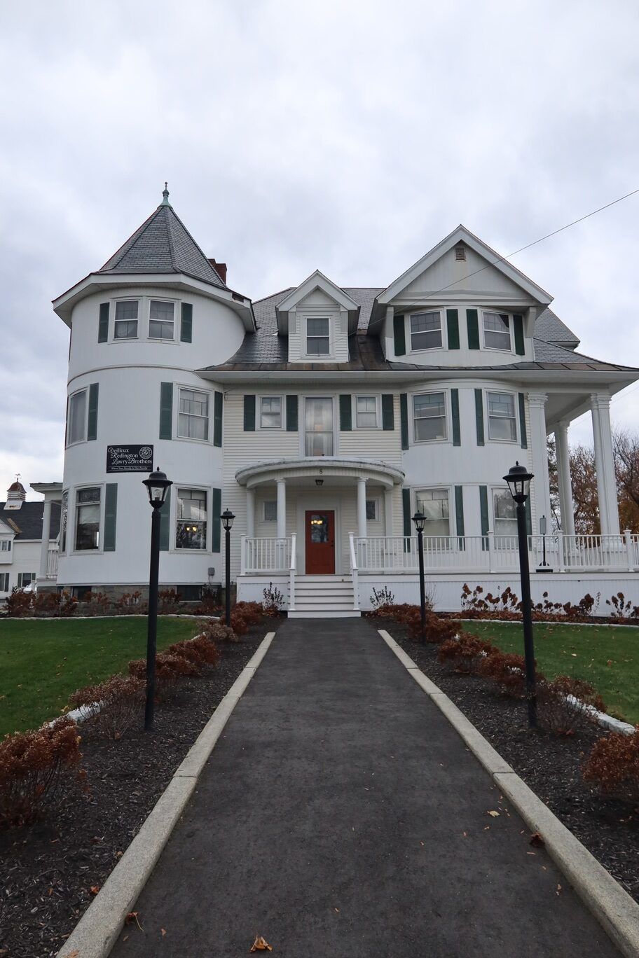 White two-story house with a tower and porch, approached by a paved pathway. Gray sky overhead.