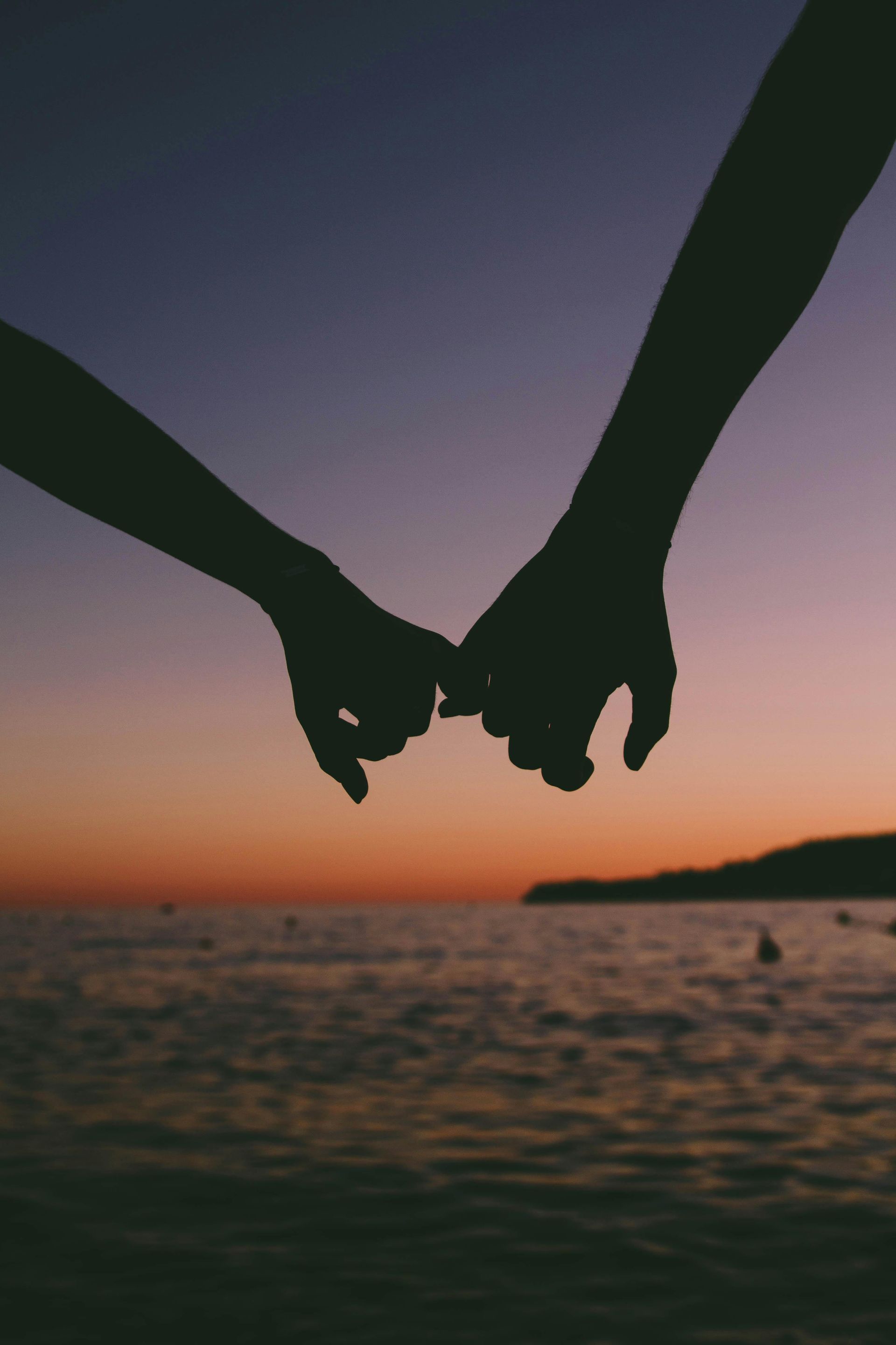 Silhouette of two hands holding pinky fingers against a sunset over the ocean.