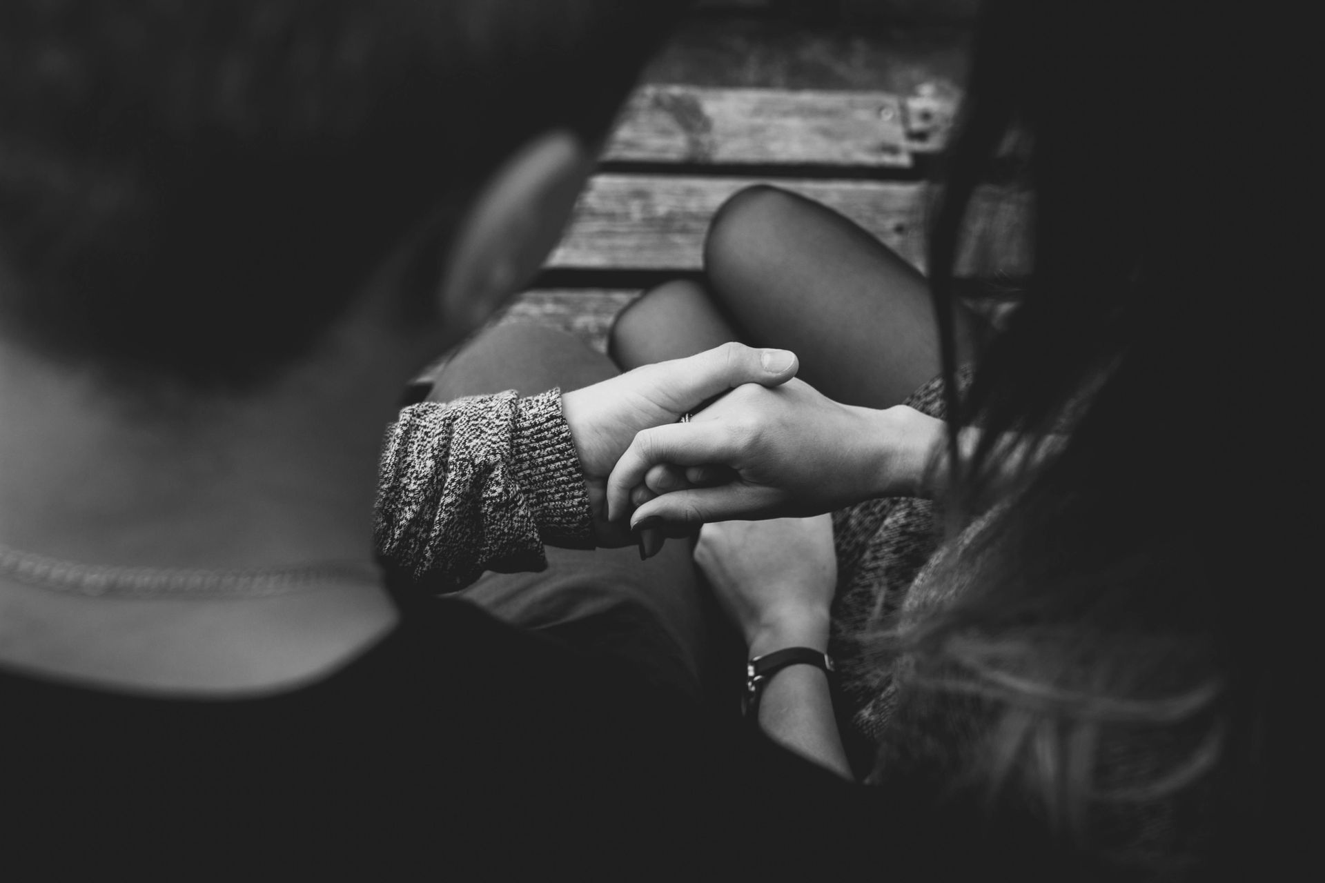 Couple holding hands, seated. Arm with patterned sleeve and necklace in view. Black and white photo.