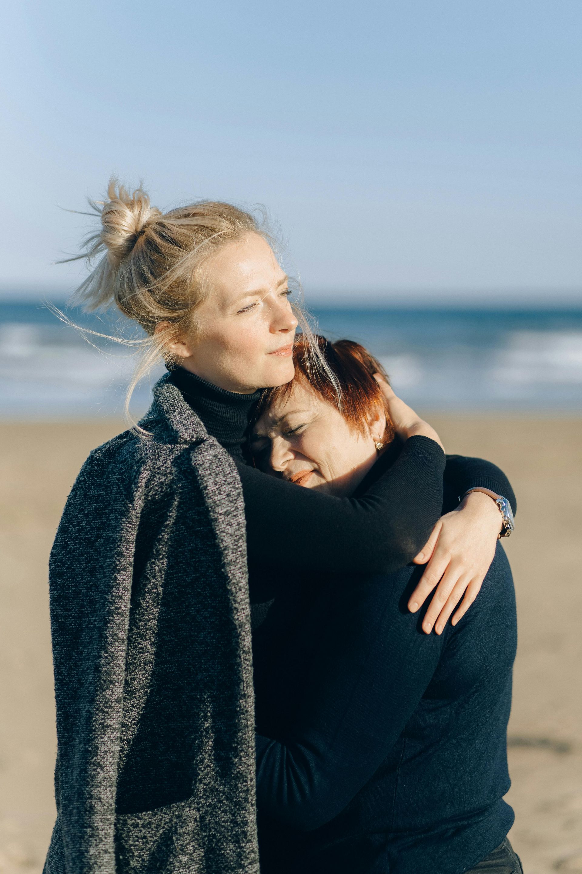 Woman with blonde bun hugs another with red hair on a beach. Both wearing dark sweaters, ocean in background.